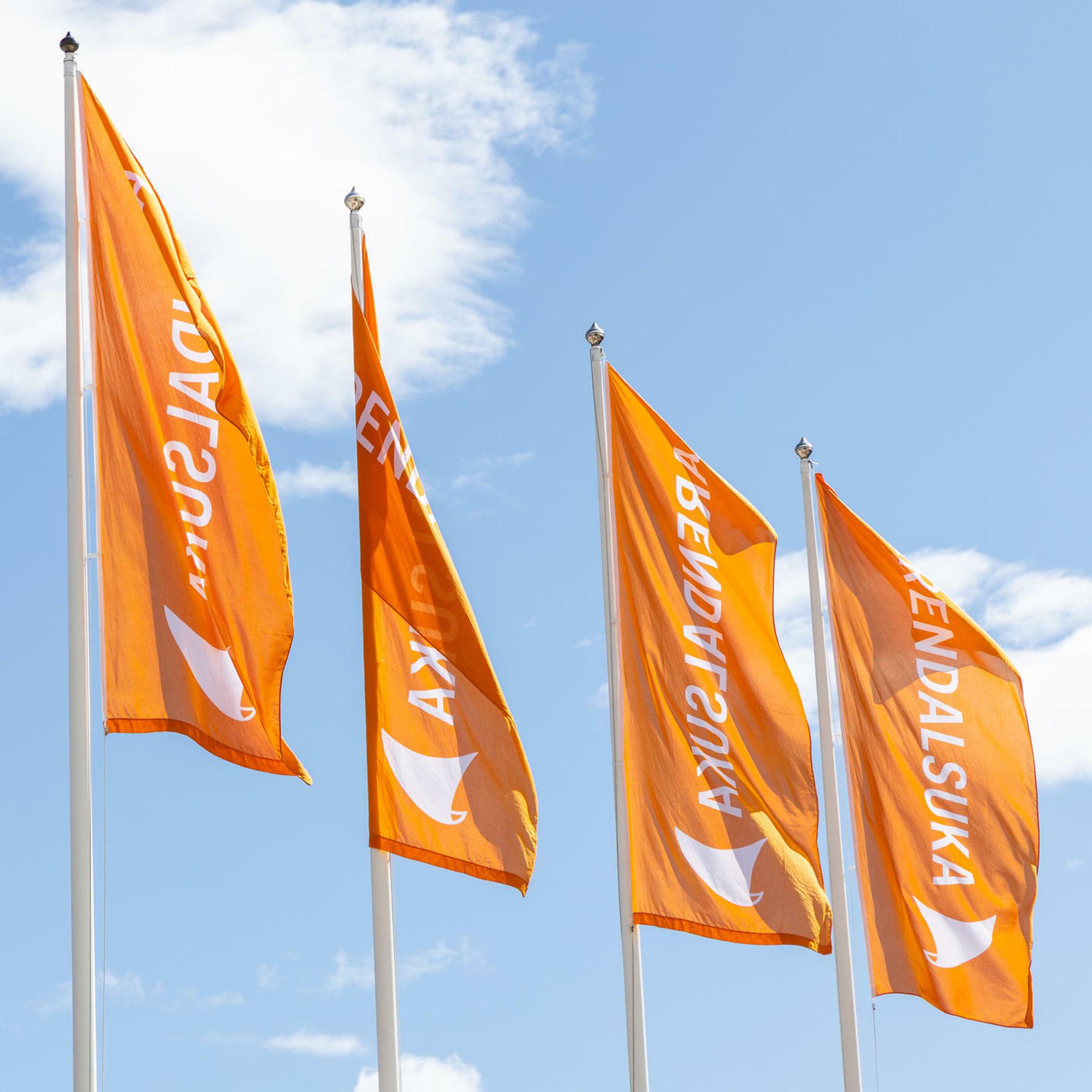 Four orange flags waving in the wind against a blue sky with white clouds. The flags display the text "ARENDALSUKA" and a logo of Arendalsuka.