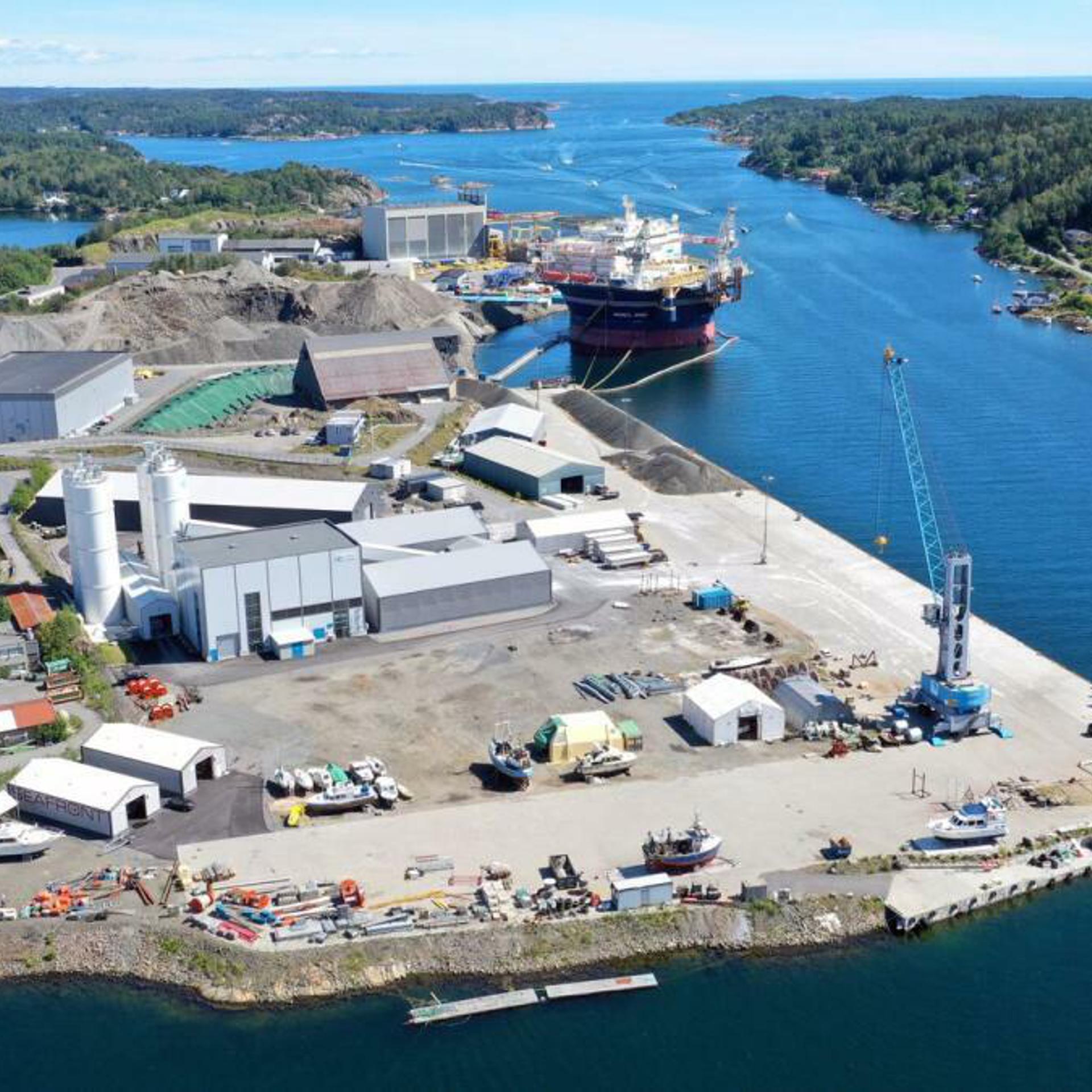 An aerial view of a Eydehavn port, showing various buildings, storage areas, and cranes. Several ships are docked at the port, with a large vessel in the central part of the image.