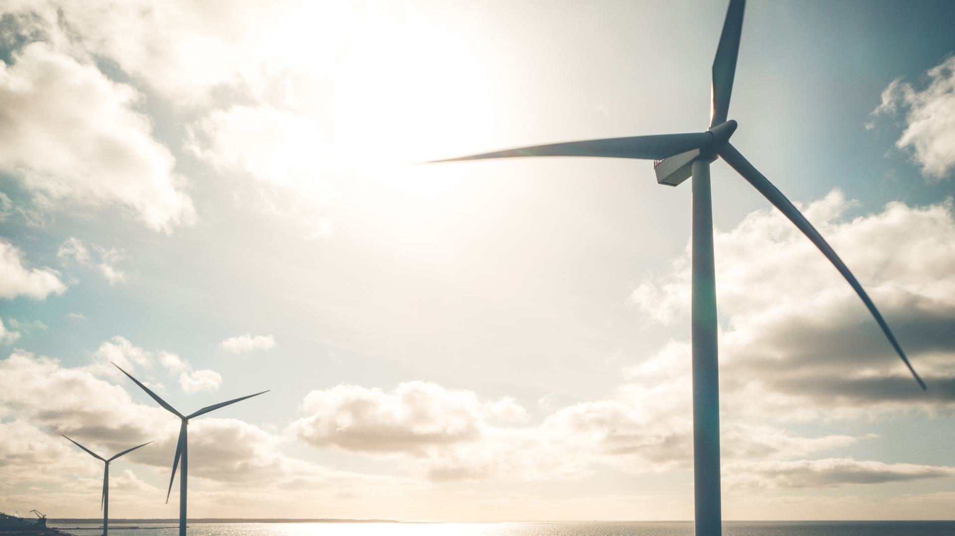 A photograph showing three large wind turbines standing in the ocean. The turbines are set against a bright, sunny sky with scattered clouds, casting reflections on the water surface below.