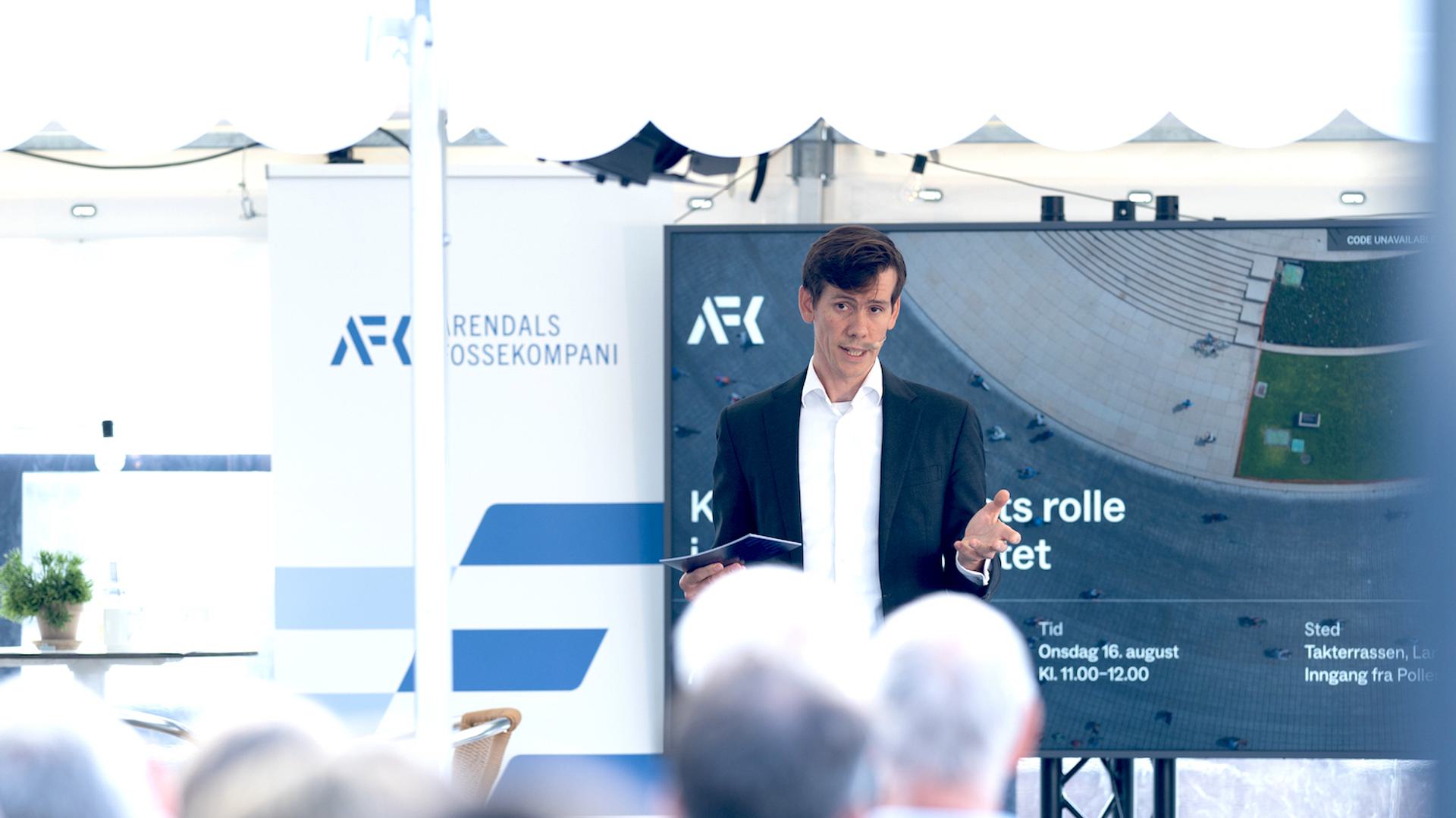 Portrait of CEO Benjamin Golding giving a presentation in front of an audience, with a screen and banner displaying "Arendals Fossekompani" in the background.