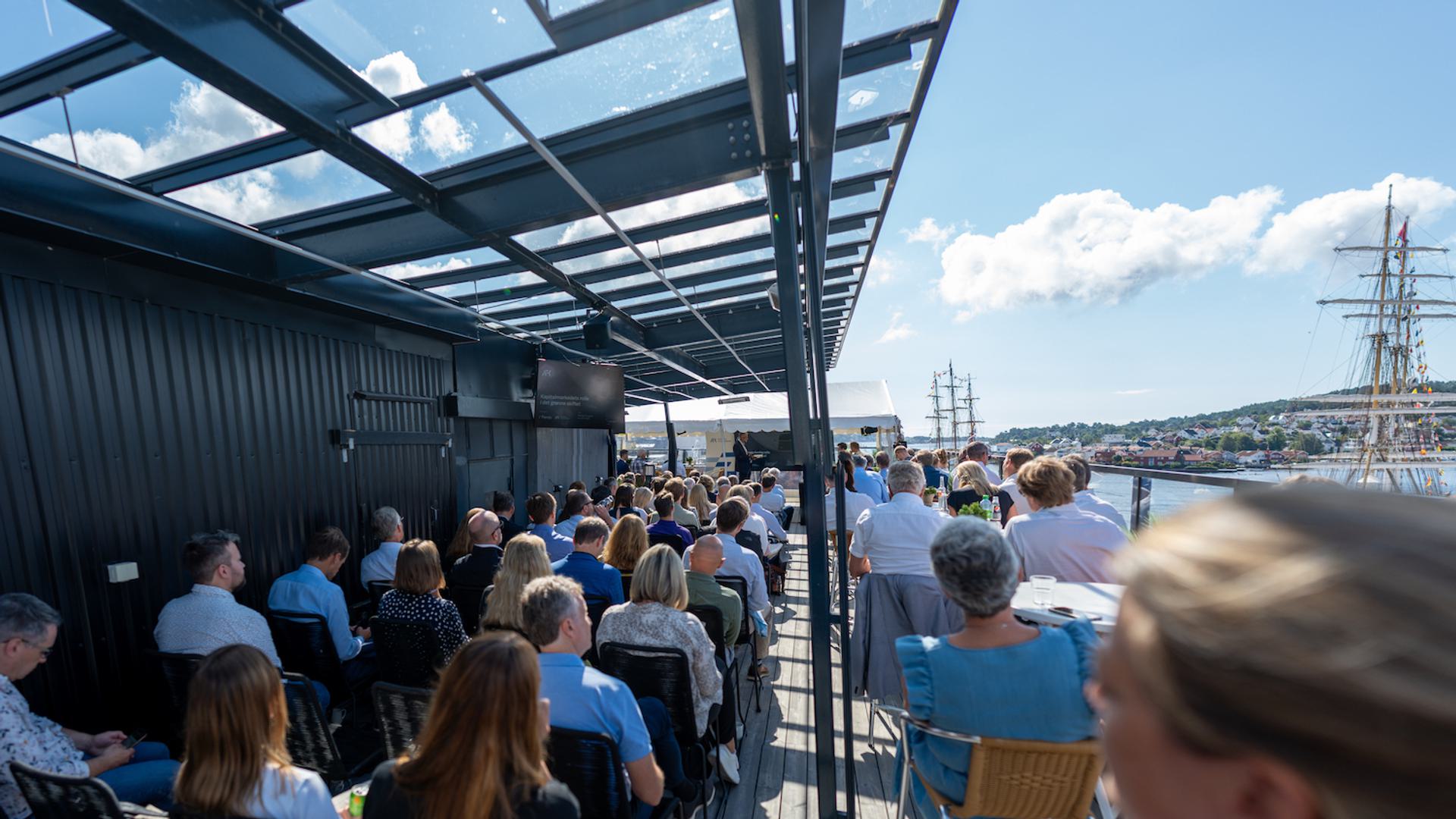 A group of people seated on the rooftop terrace at Arendals Fossekompani, attending the event. The view includes a waterfront with sailboats and buildings in the background.