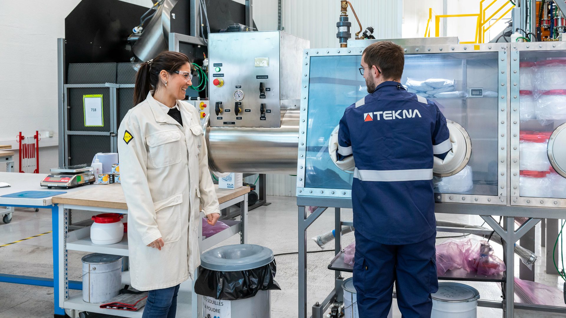 Two employees working in an industrial lab setting. The person on the left is a woman wearing a white lab coat, standing and smiling. The person on the right is a man wearing a blue uniform with "TEKNA" on the back, working with equipment inside a controlled environment cabinet.