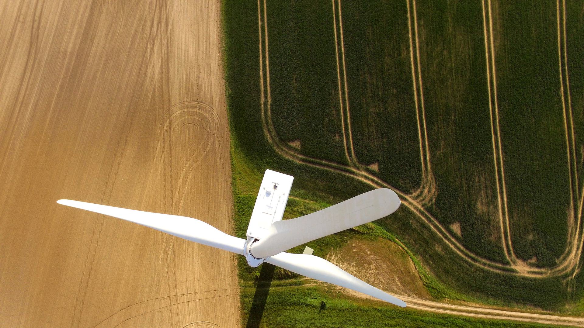 An aerial view of a wind turbine situated in a field. The turbine's blades extend over a landscape that includes patches of green grass and brown soil.
